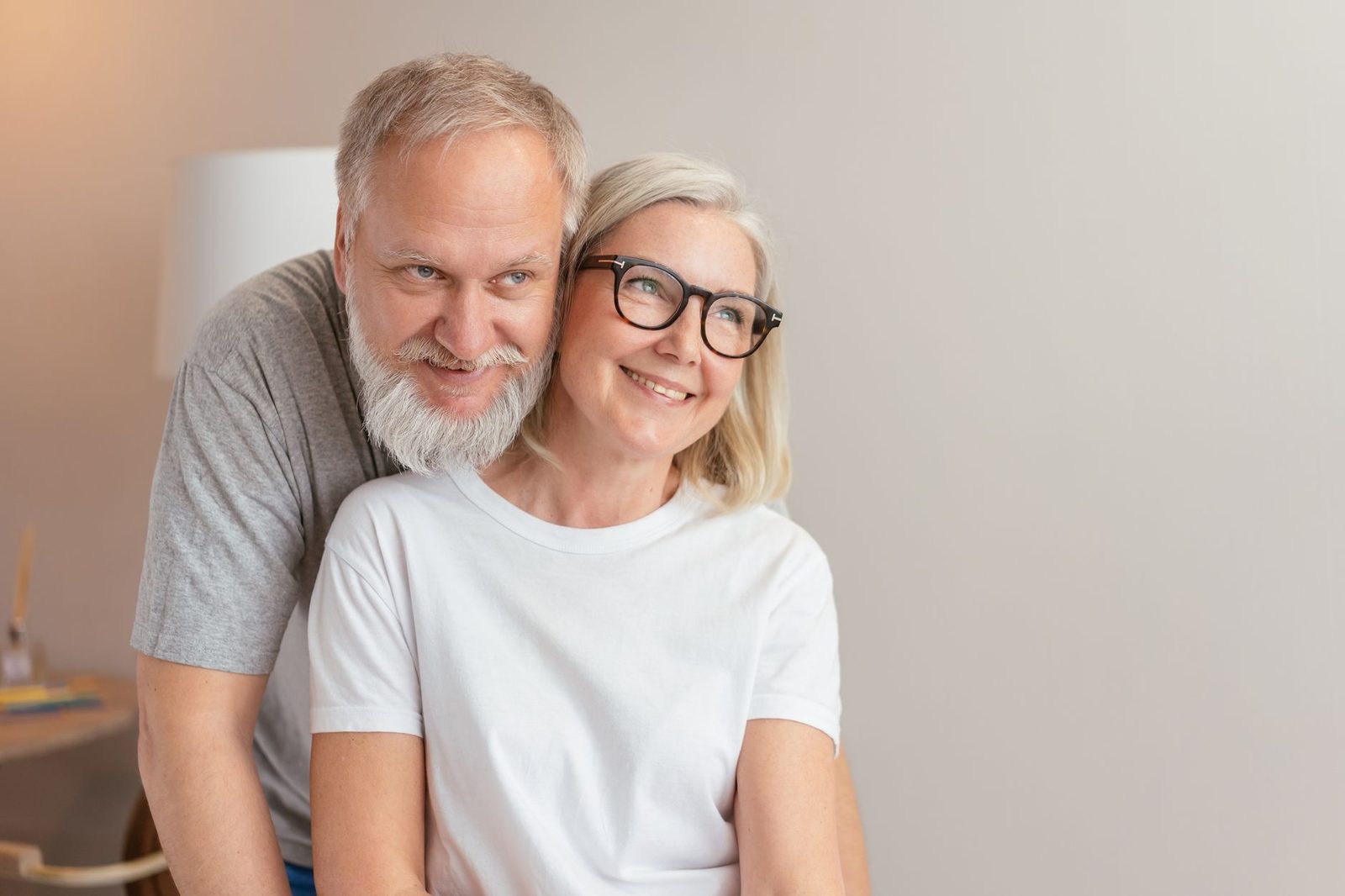 Casal idoso sorridente compartilhando um abraço amoroso dentro de casa, ambos usando óculos