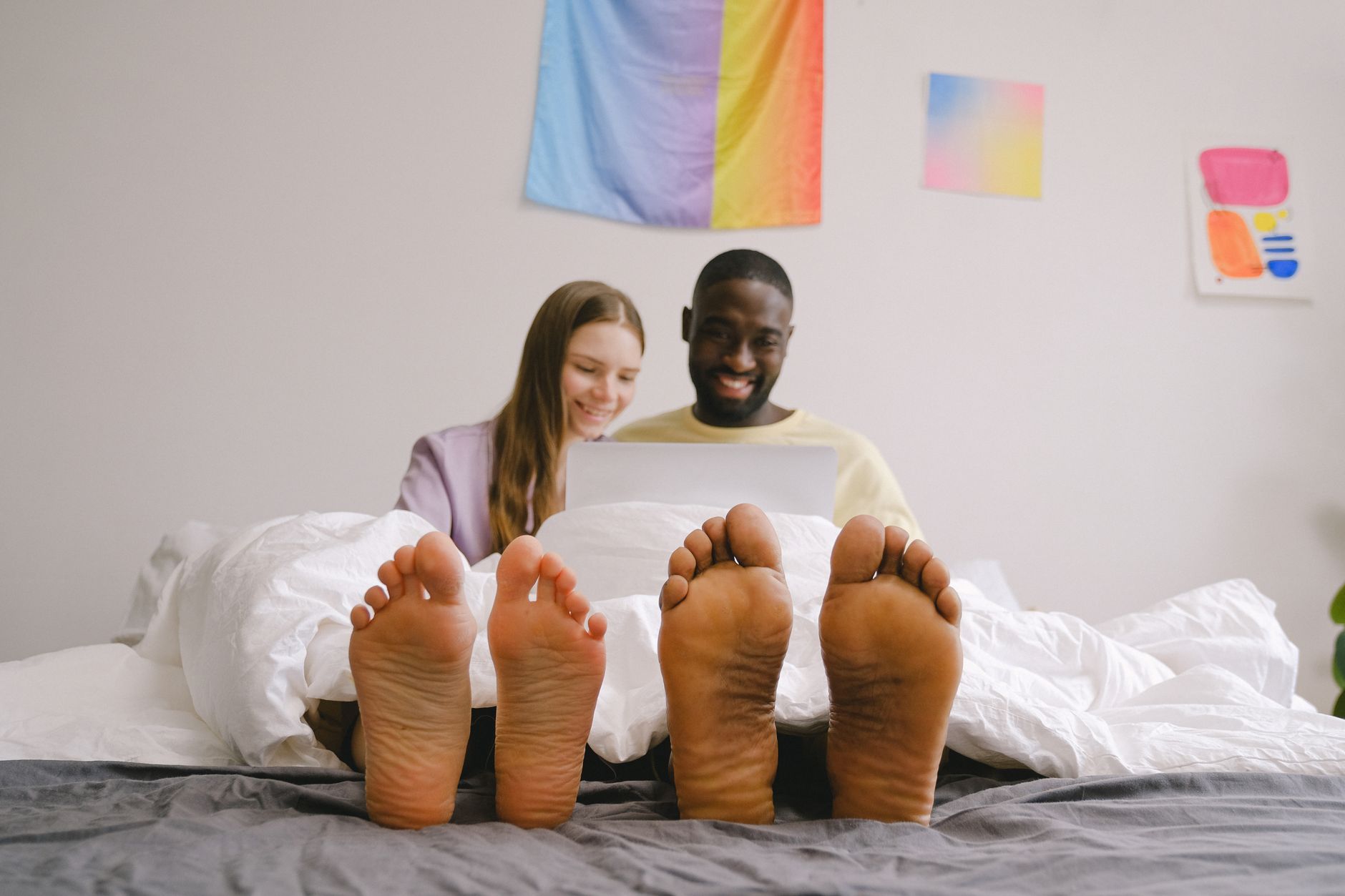 Casal interracial se conectando em ambiente interno, relaxando sob a coberta com laptop, aproveitando um tempo casual juntos