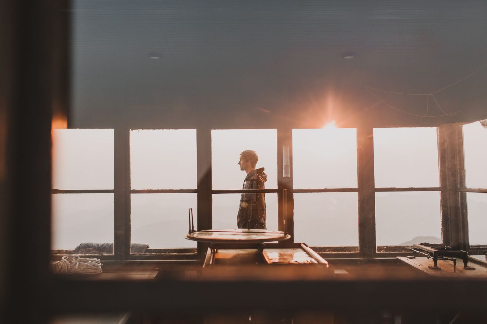 Um homem contemplativo está em um quarto iluminado pelo sol com grandes janelas de vidro, capturando um momento sereno
