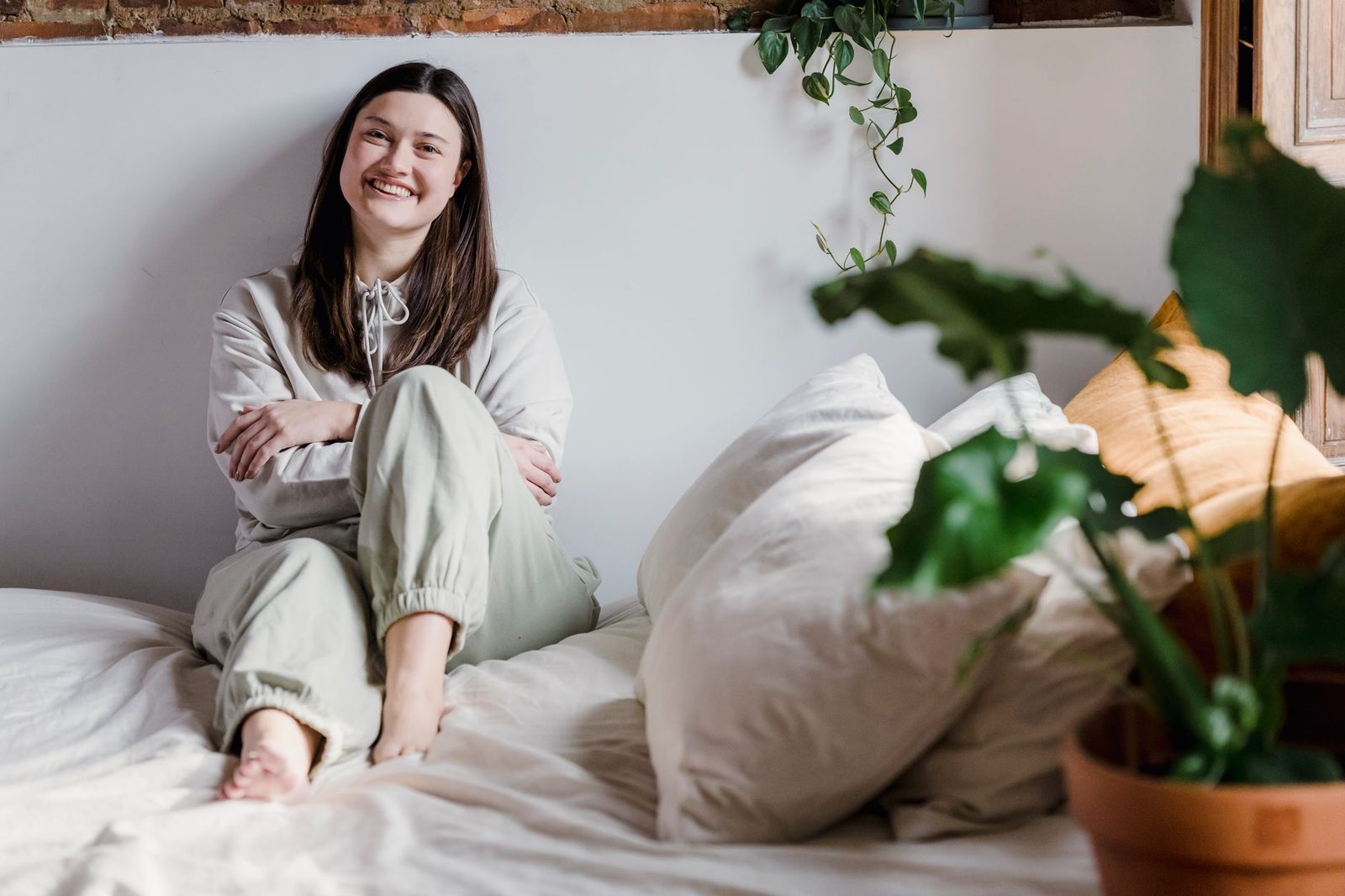 Uma mulher alegre senta-se confortavelmente em uma cama em um quarto aconchegante, cercada por almofadas e plantas