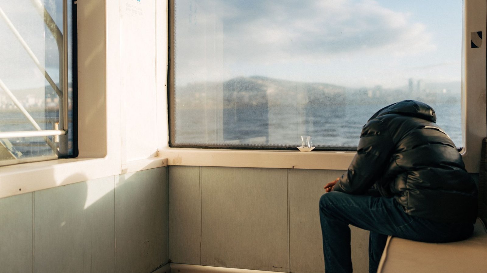 Um homem com um paletó descansa em um ferry em Istambul, olhando para o mar através de grandes janelas