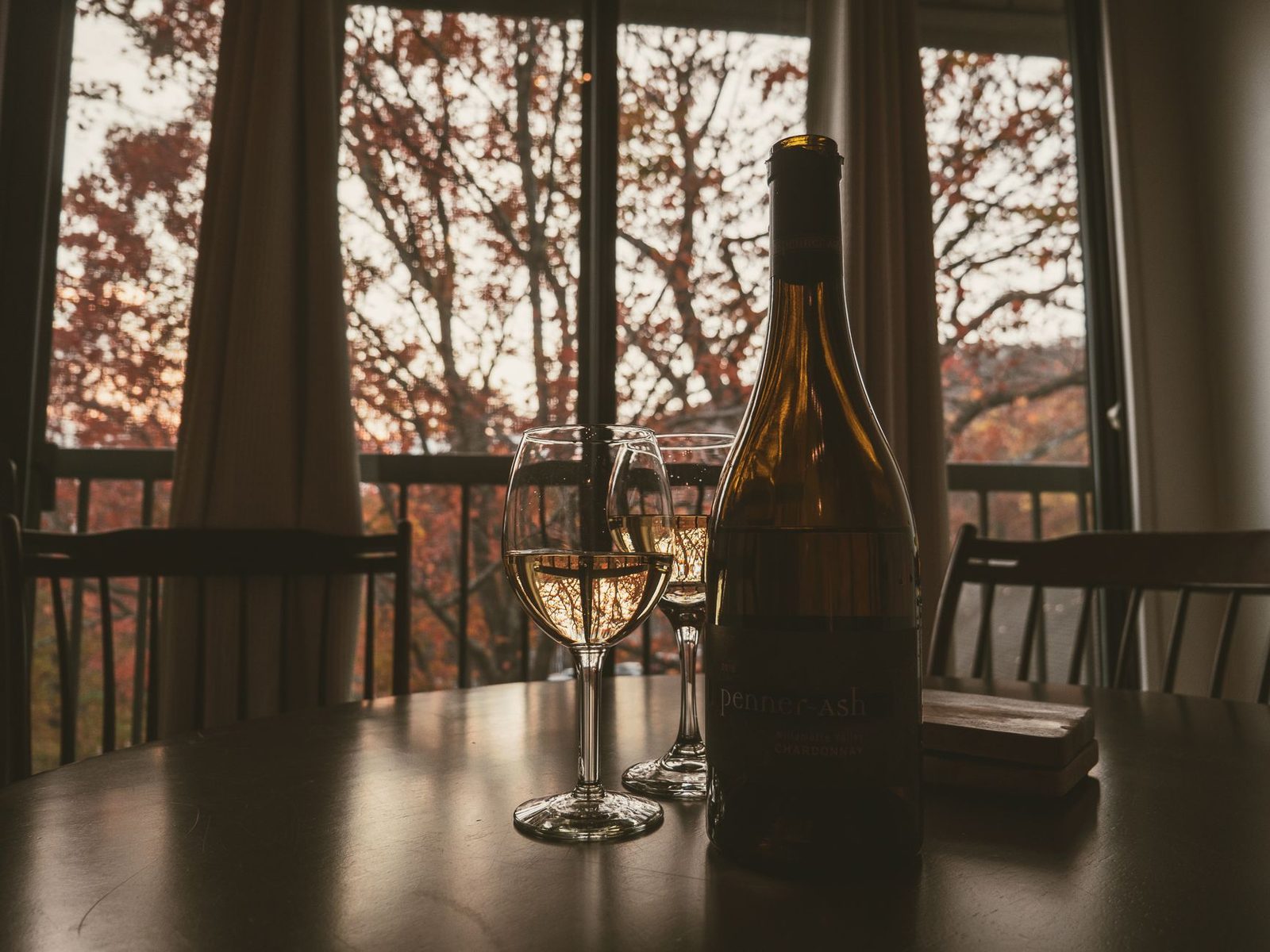Um ambiente quente e aconchegante com dois copos de vinho branco e uma garrafa no interior, emoldurado por árvores de outono