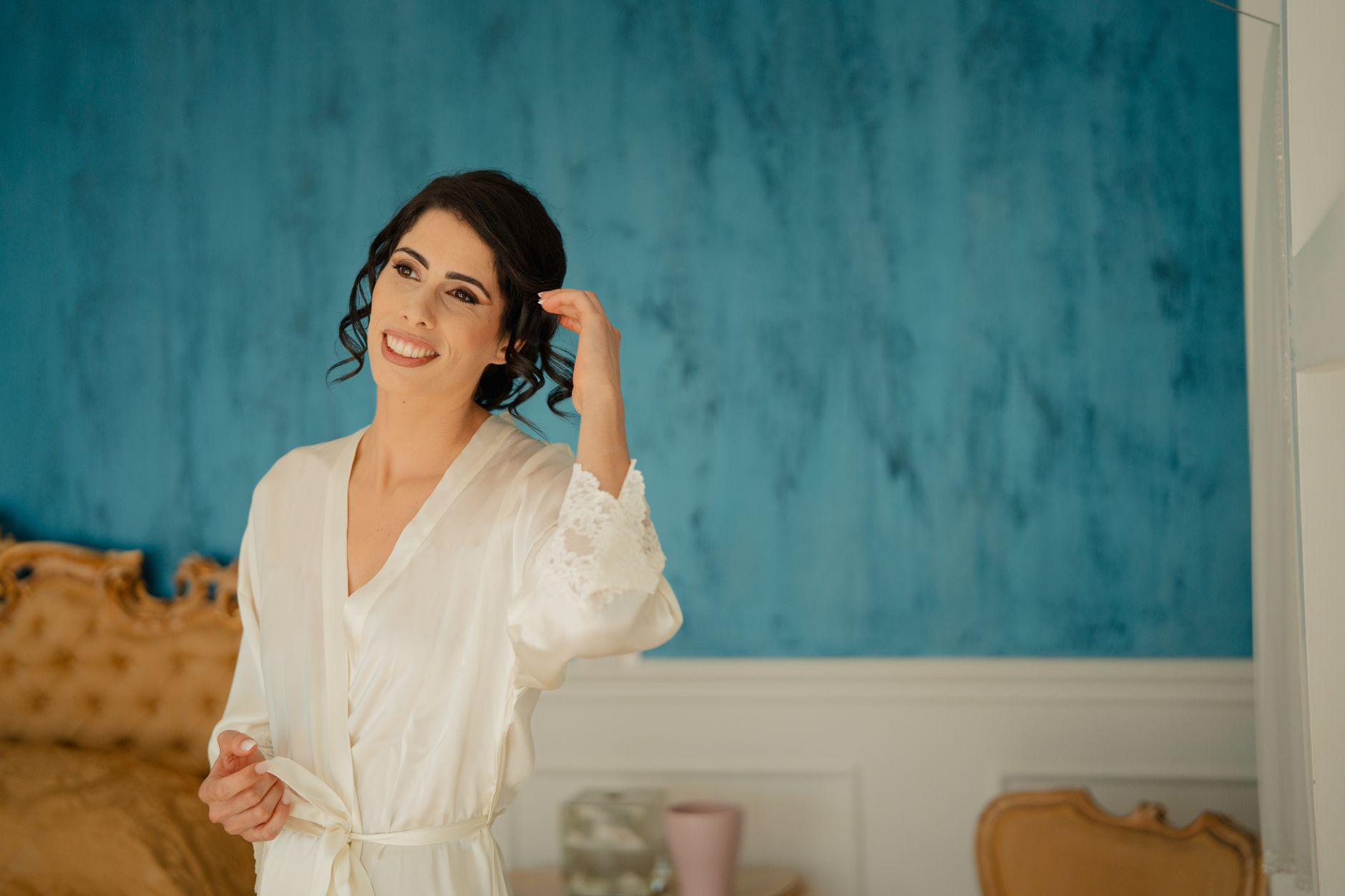 Noiva sorridente de robe branco de cetim com renda, se arrumando no quarto