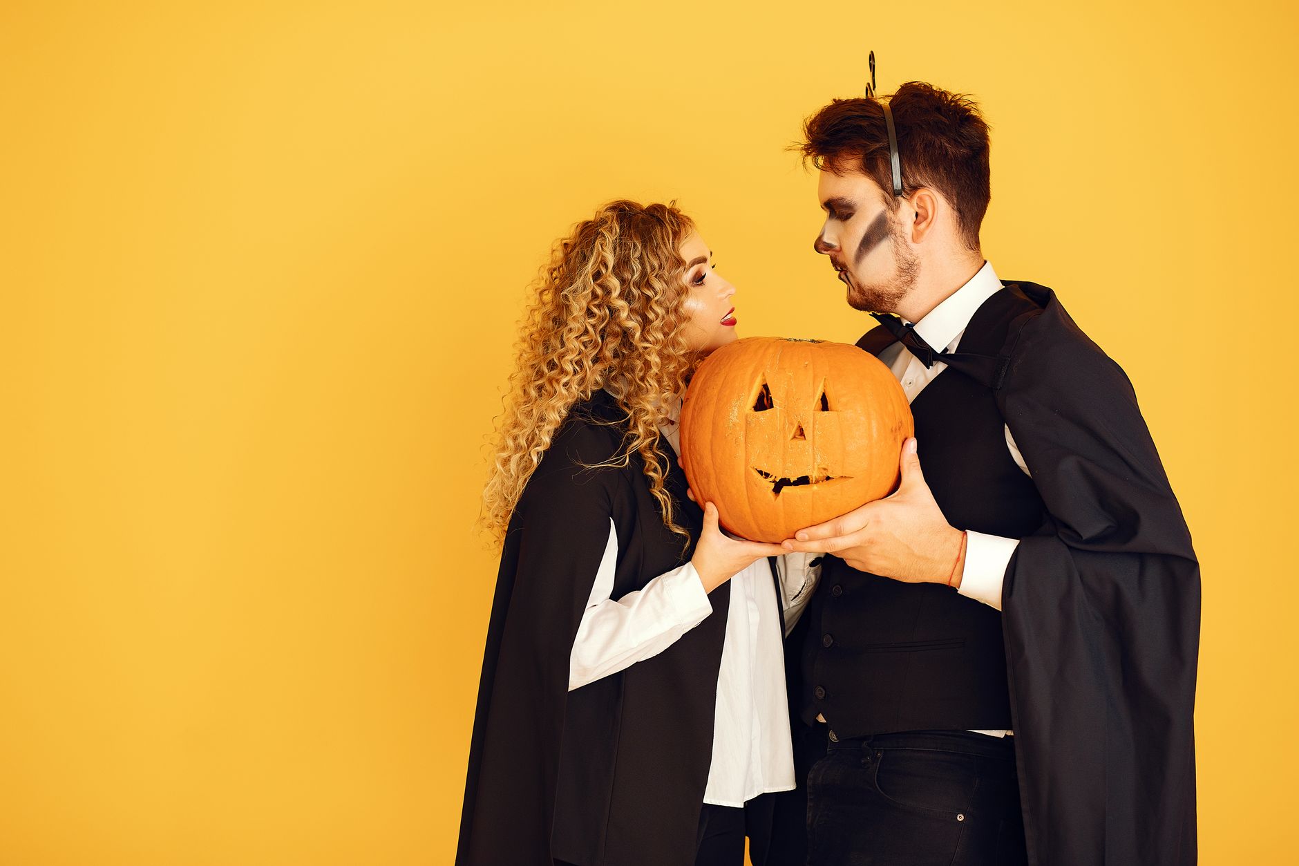 Casal fantasiado para o Halloween, com capas pretas e maquiagem, segurando juntos uma abóbora esculpida