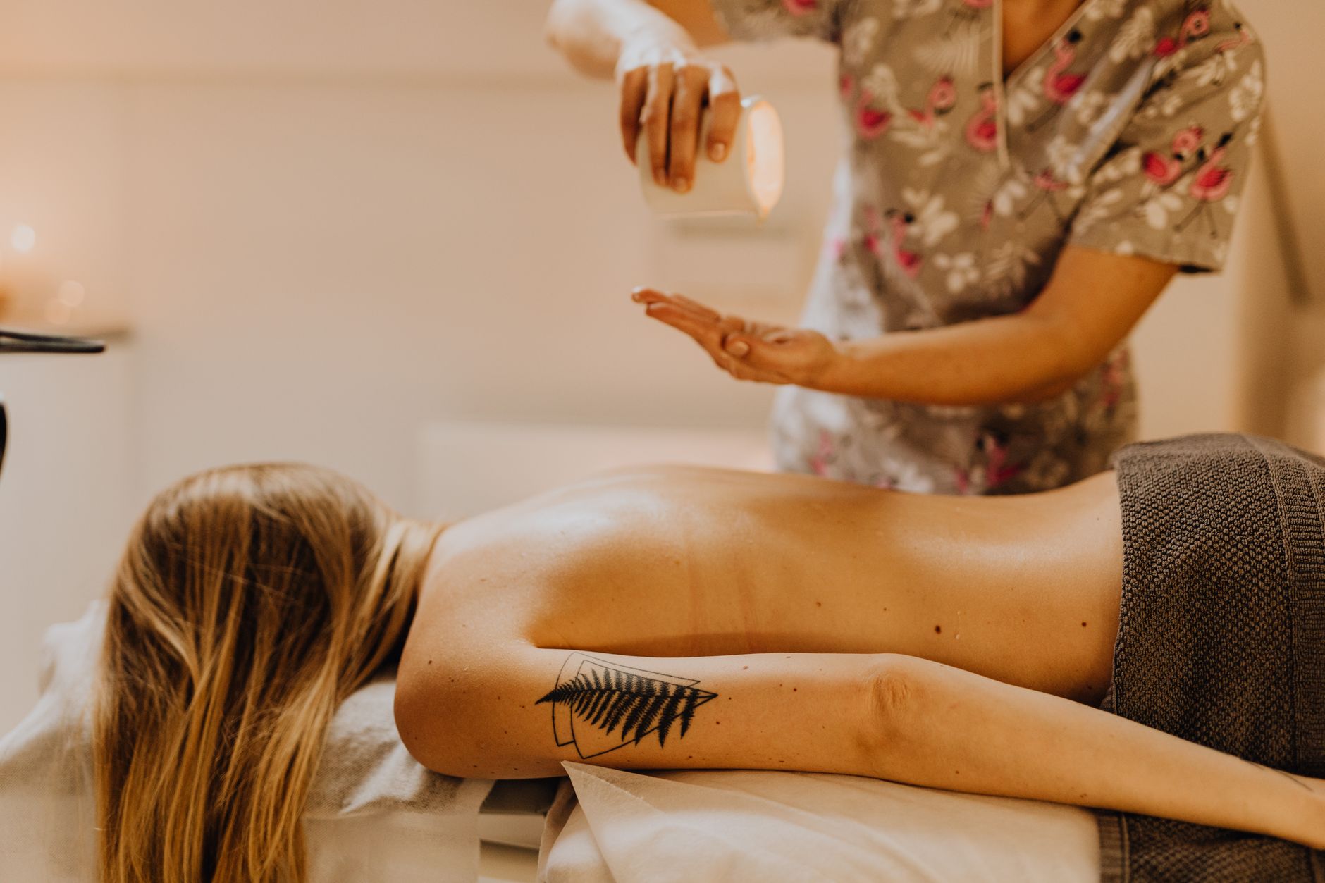 Mãos despejando óleo de massagem morno sobre as costas de uma mulher deitada, em ambiente com luz suave
